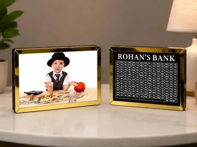 Two gold-framed money box banks displayed on a white table. The left bank shows a custom photo of a young boy in a hat, and the right bank displays a black account summary panel with the text &