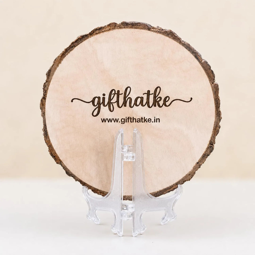 A round natural wood slice coaster with a rustic bark edge, displayed on a clear acrylic stand. The top of the wood is printed with the brand name &